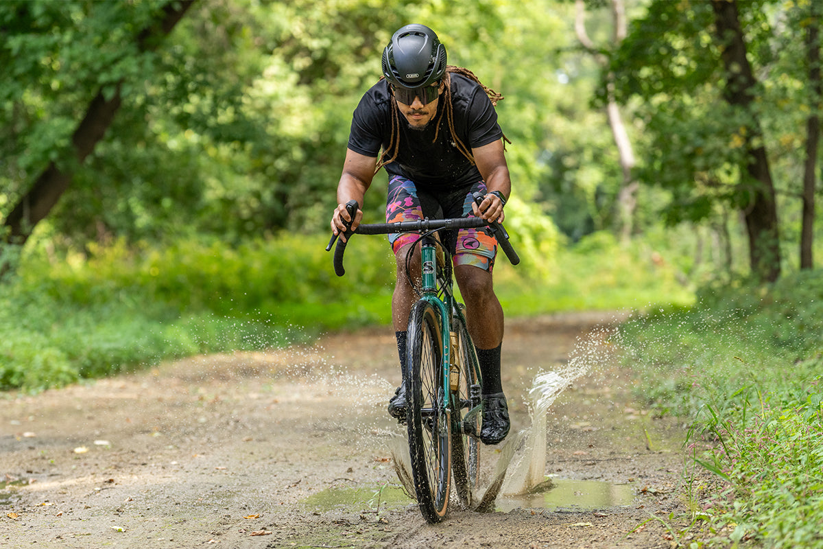 Cyclist riding Surly Straggler bike in Subtropic Algae color on gravel road splashing through puddle