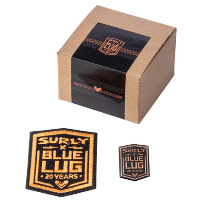 A brown cardboard gift box with a black wrap label reading 'SURLY x BLUE LUG' is shown above two embroidered patch designs. The larger patch reads 'SURLY x BLUE LUG 20 YEARS' in tan and black, and the smaller one is a smaller matching patch.