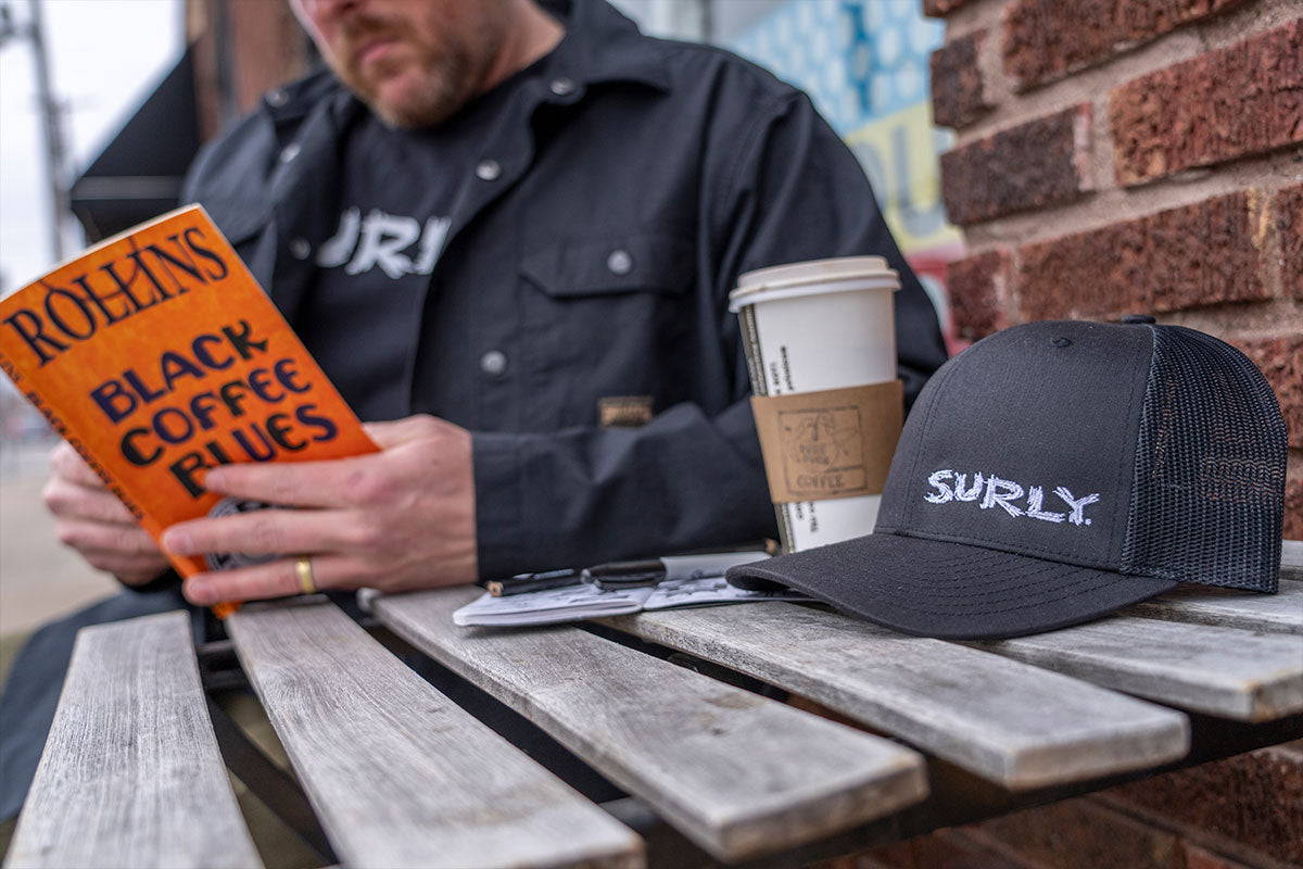 Person reading book outside with Surly hat and coffee on table