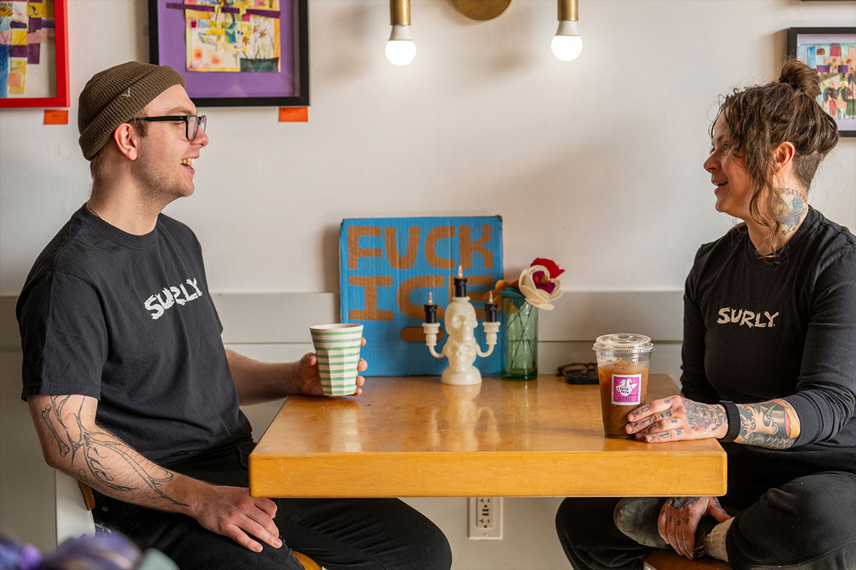 Two people sitting at a table with drinks, wearing Surly shirts, in a casual setting.