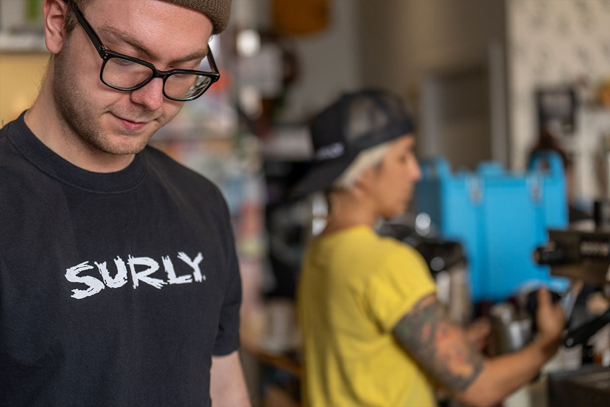 Person wearing a 'Surly' t-shirt in a casual setting with another person in the background.