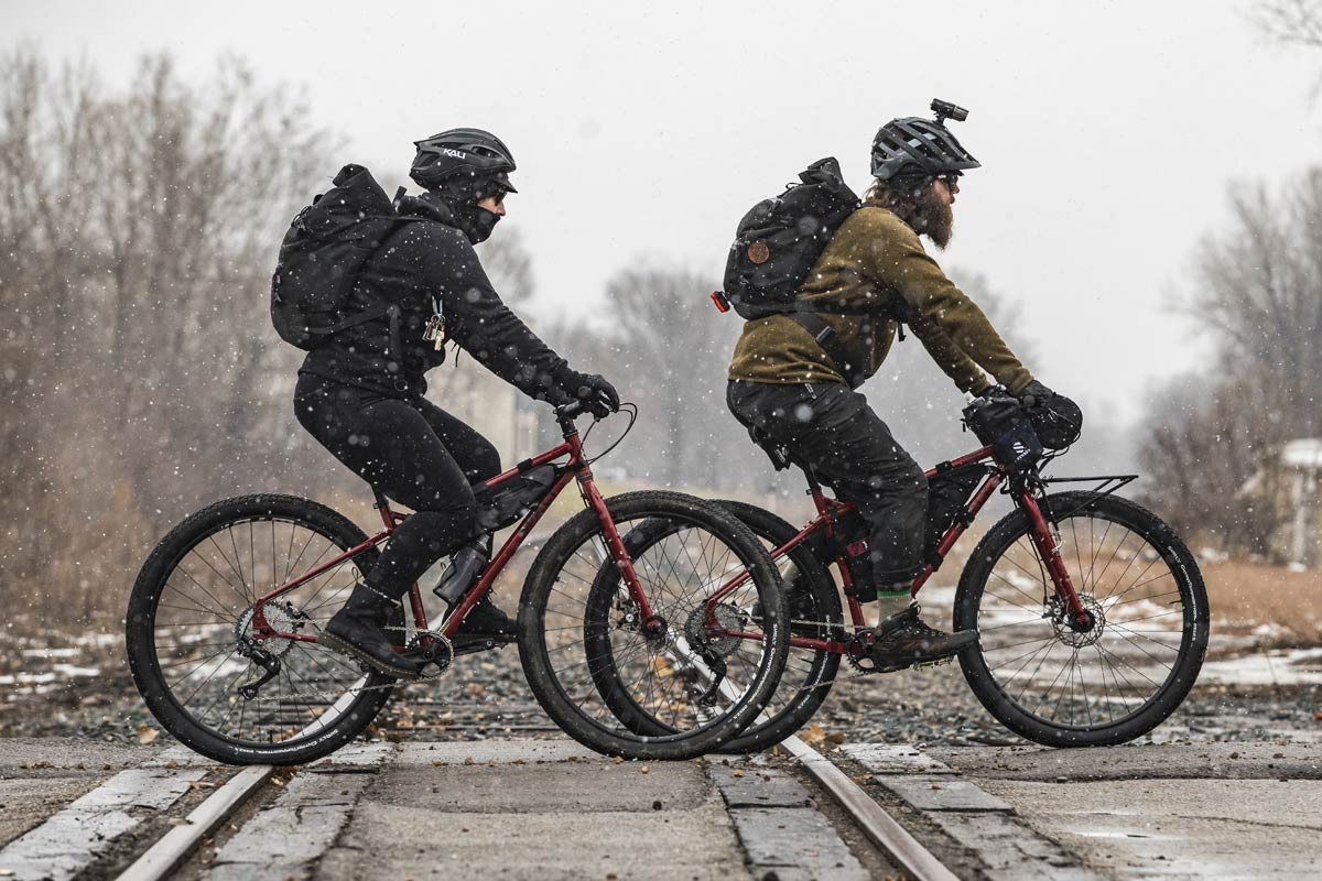 Two cyclists riding Ogre bikes on road across train tracks wearing warm clothing, snow falling