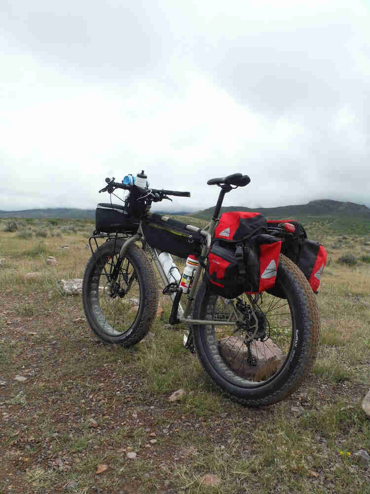 Rear, left side view of a tan Surly fat bike, loaded with gear, on a rocky grass field with hills ahead in the distance