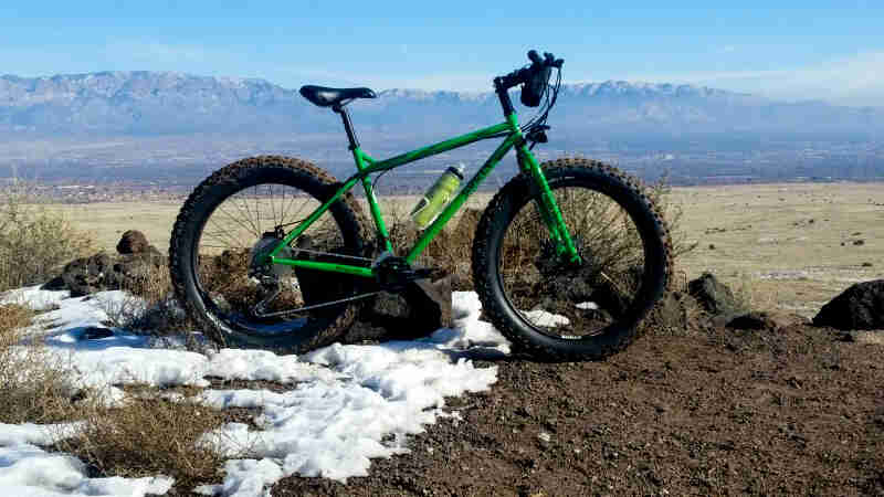 Right side view of a green Surly fat bike, parked on a snow patch and gravel, with mountains in the background