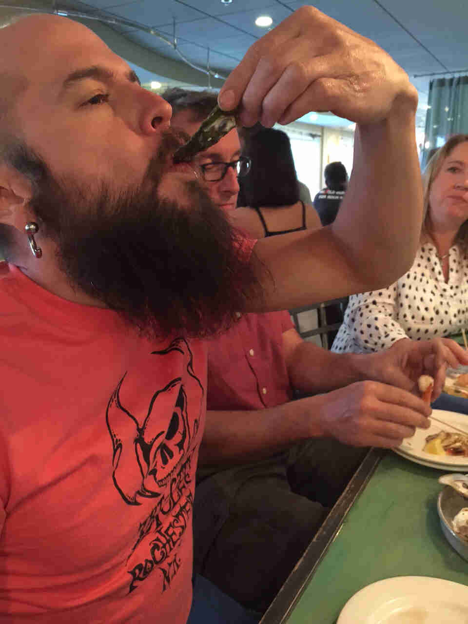 Right side view of a person sliding an oyster into their mouth from the shell, with people sitting behind them watching
