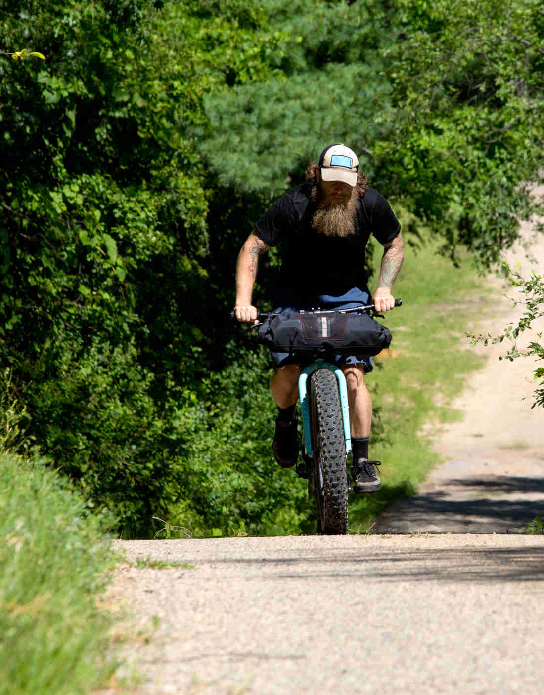 Front view of a cyclist on a light blue Surly fat bike, right up a hill on a gravel road, with trees in the background