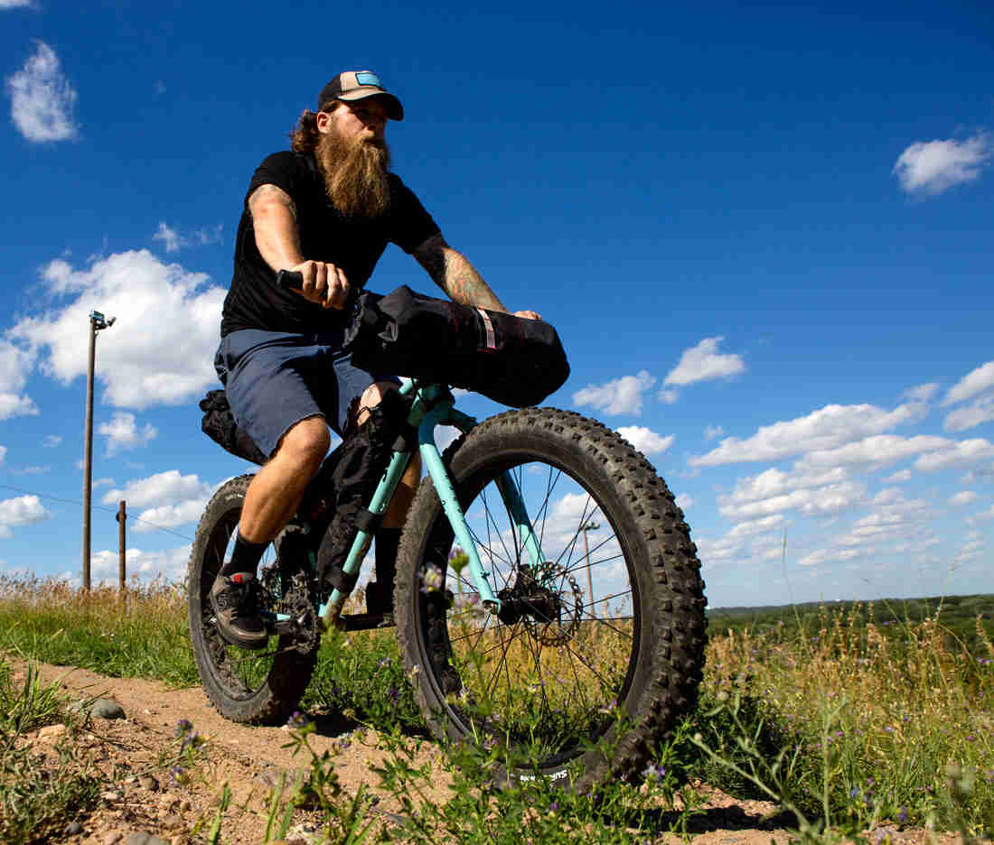 Upward, right side view of a cyclist riding a light blue Surly fat bike with gear, on a dirt trail in a grass field