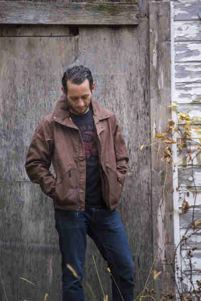 Front view of a person wearing a Surly V2 Waxed canvass jacket, brown, in front of a wood building