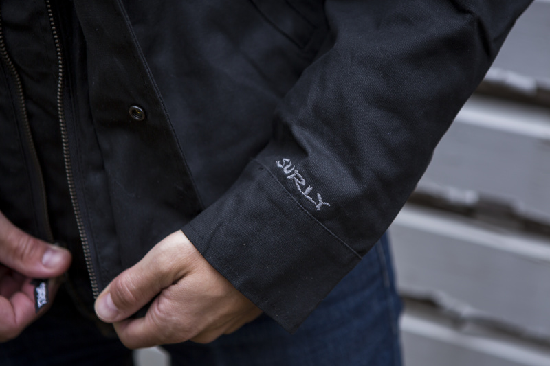 Cropped view of the Surly embroidered sleeve and zipper wind flap, on a V2 Waxed canvass jacket, black