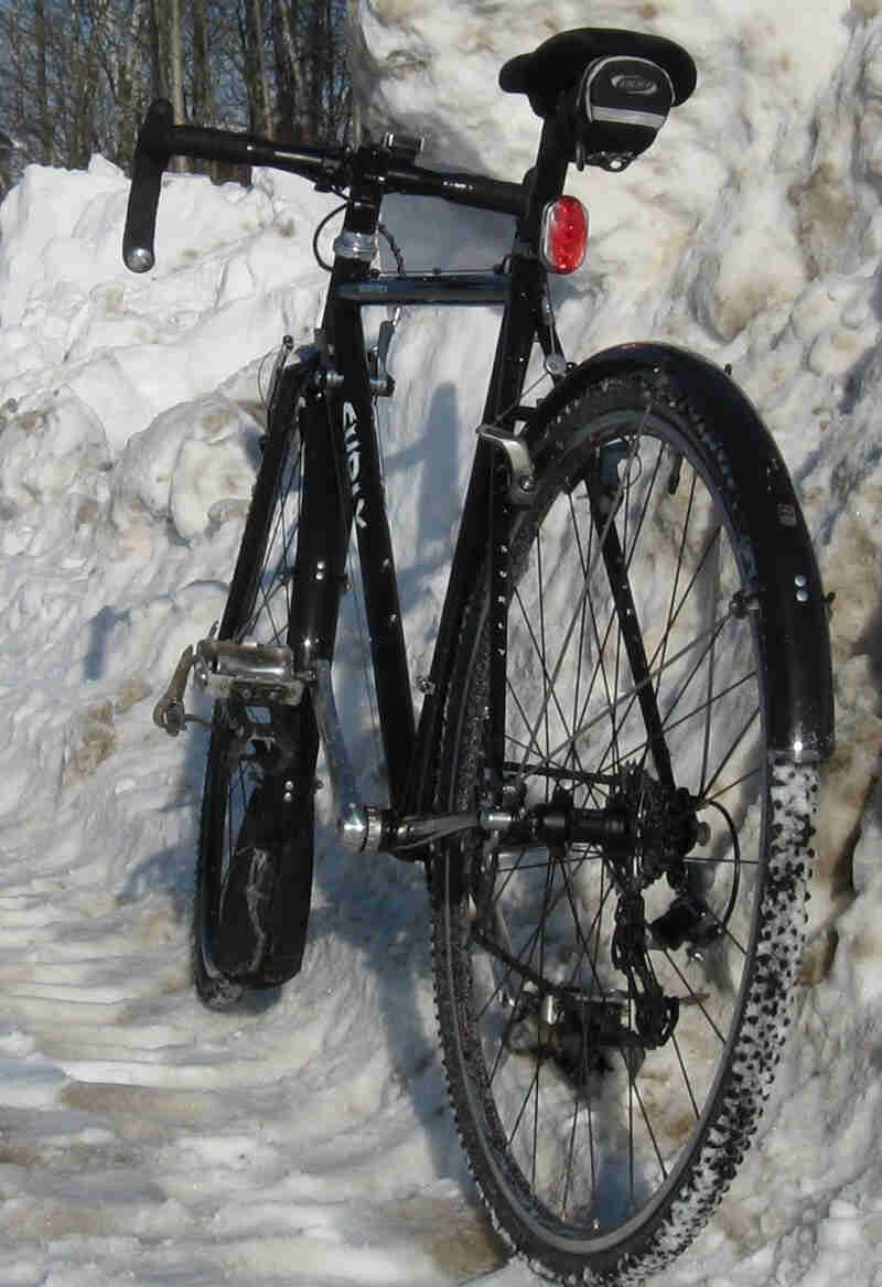 Rear, left side view of a black Surly bike with fenders, leaning against a tall snowbank, near the woods