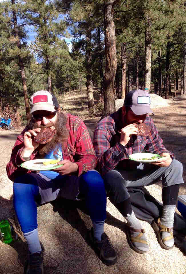 2 People wearing baseball caps, sit side by side on a rock eating breakfast, with the forest in the background