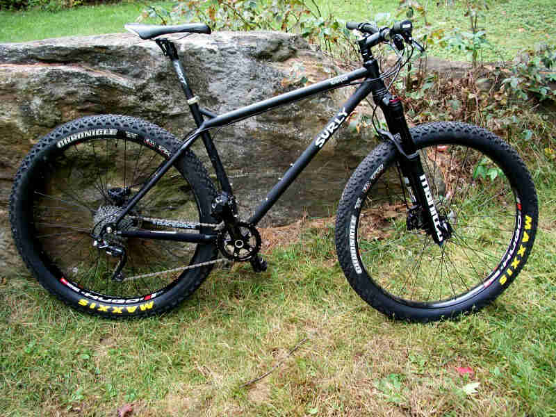 Right side view of a Surly Krampus bike, matte black, parked on grass in front of a rock wall