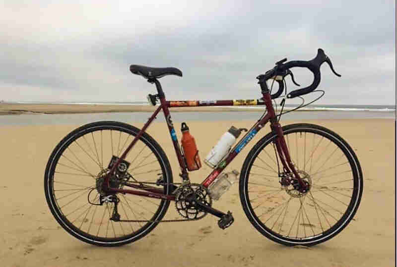 Right side view of a red bike with 2 water bottles,  parked on a flat beach, with the ocean in the background