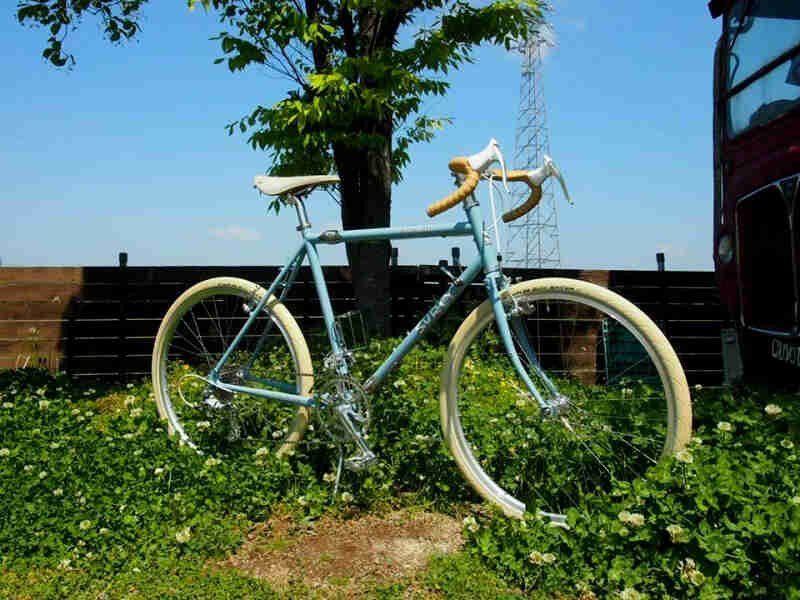 Right side view of a mint Surly bike with white tires, parked in flowers, with trees and a wood fence in the background