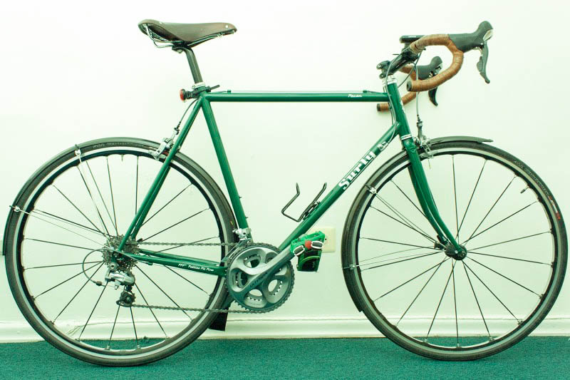 Right side view of a green Surly Pacer bike with fenders, parked along a white wall in a room