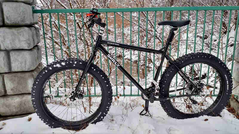 Left profile of a black Surly fat bike, on a snowy bridge over a stream, with trees in the background
