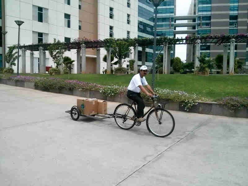 Right side view of a cyclist riding a Surly bike with a trailer hitched to the back, with buildings in the background