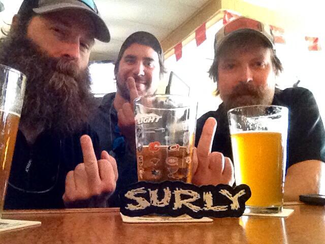 Front view of 3 people sitting at a table with beers, with a Surly Bikes patch leaning on the middle glass
