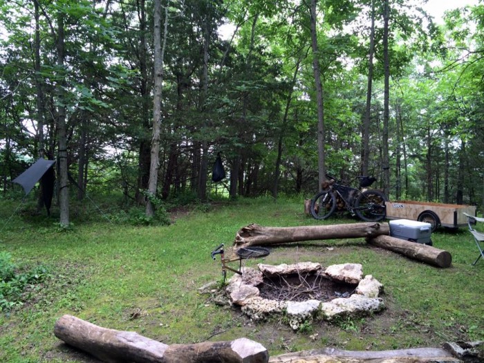 A campfire ring with stones and logs laying around it, with a bike and trailer behind, at a campsite in the woods