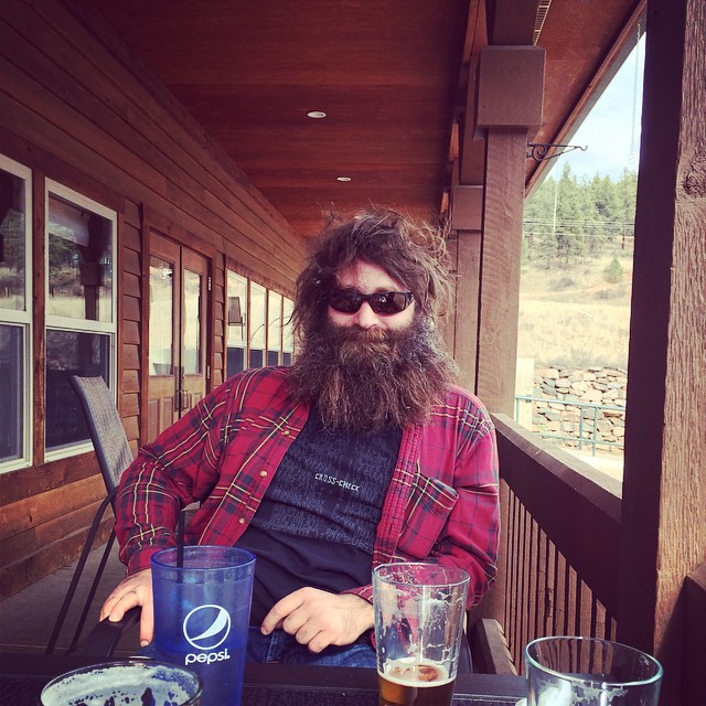 A person with a bushy beard, wearing sunglasses, sitting at a table on the porch of a wood building