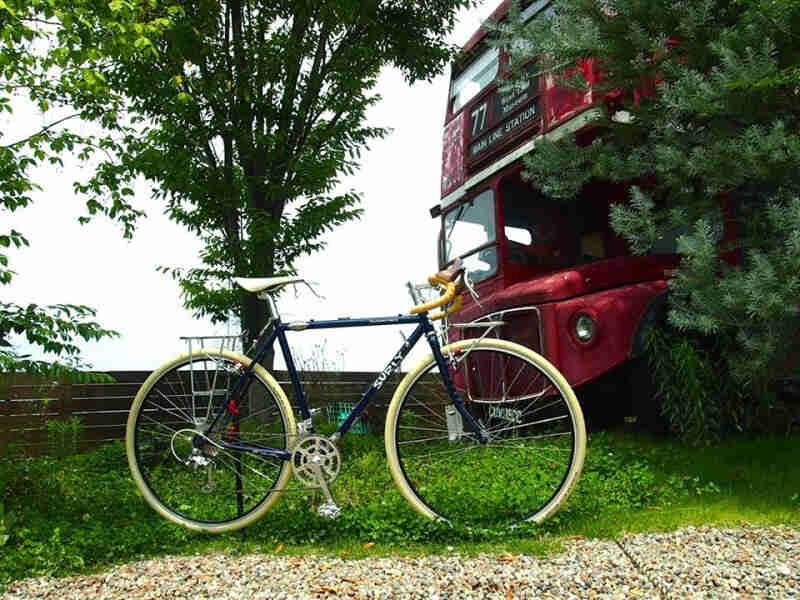 Right side view of a blue Surly bike, parked in grass in front of a double decker bus, peeking out from a tree