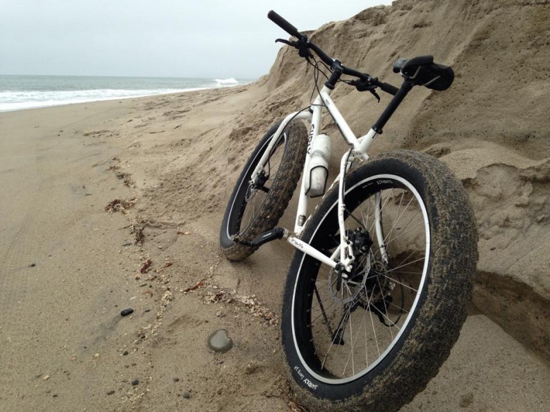 Rear, left side view of a white Surly Pugsley fat bike, leaning on a sand bank at a beach, with the ocean in background