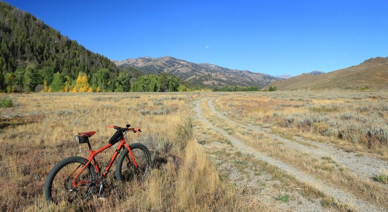 Rear, right side view of a red Surly bike, on a grassy field, next to a gravel road with tree covered hills ahead