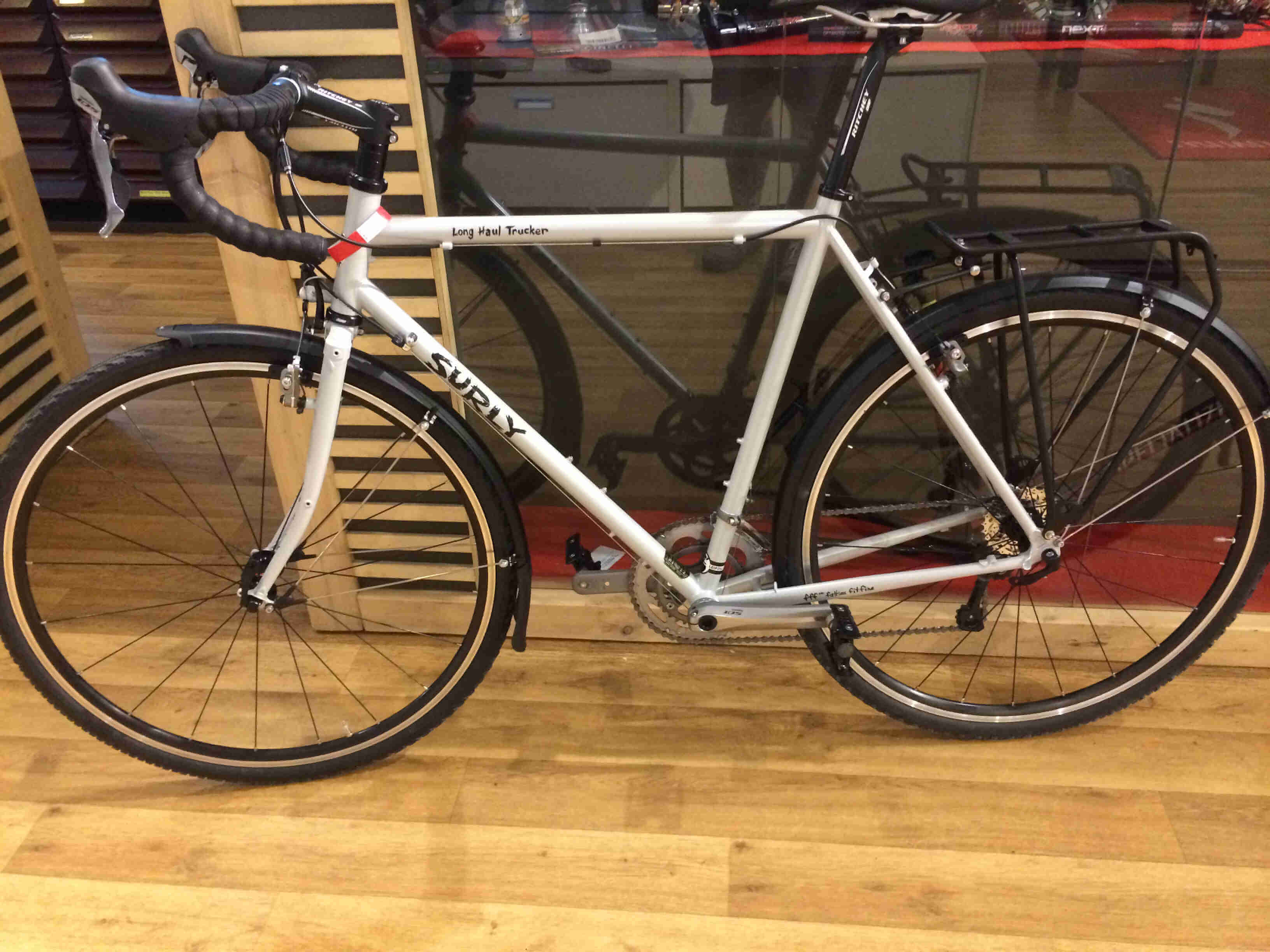Left side view of a white Surly Long Haul Trucker bike, leaning against side of a glass store counter, on wood floors