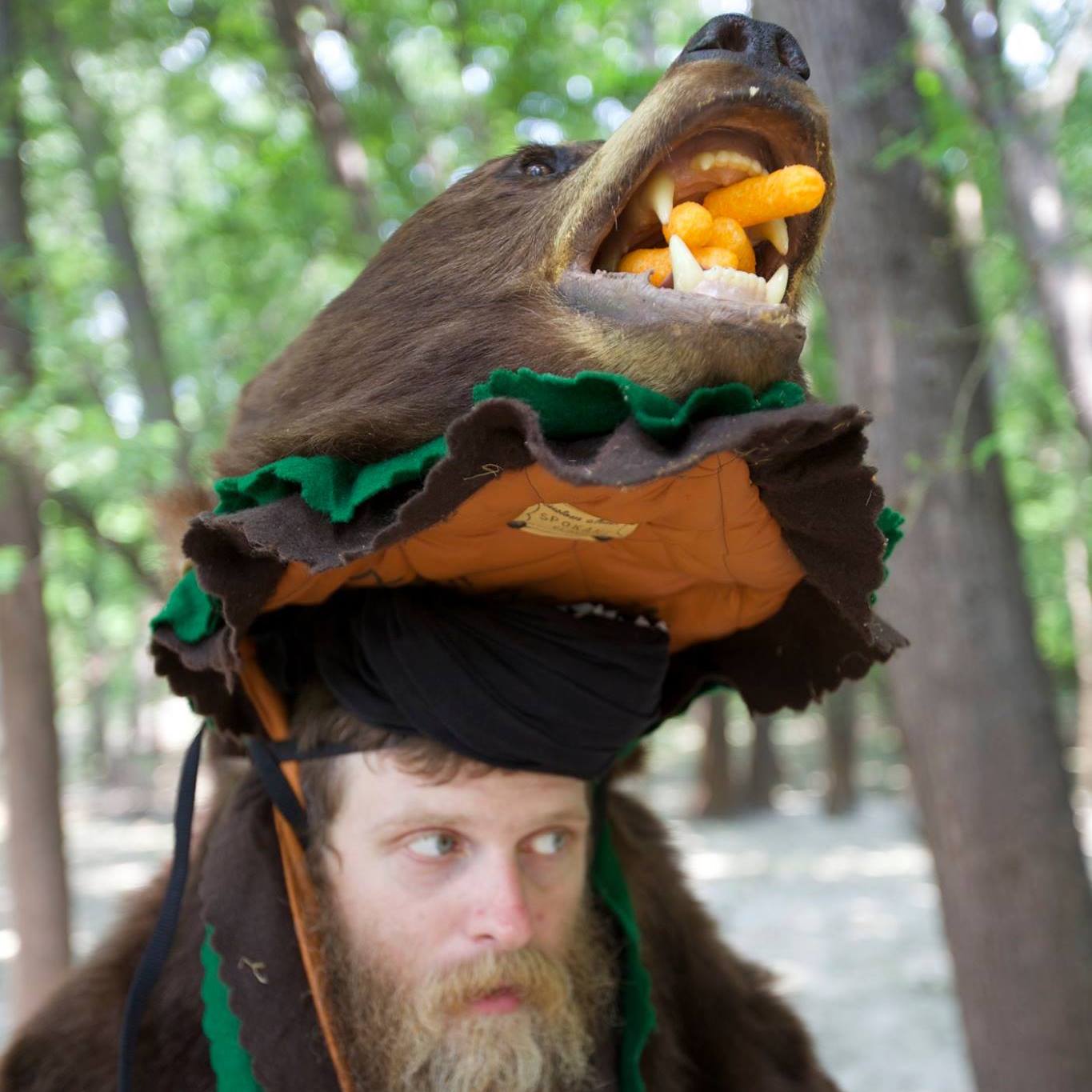 A person wearing a bear head hat with cheese puffs in the mouth, with trees in the background