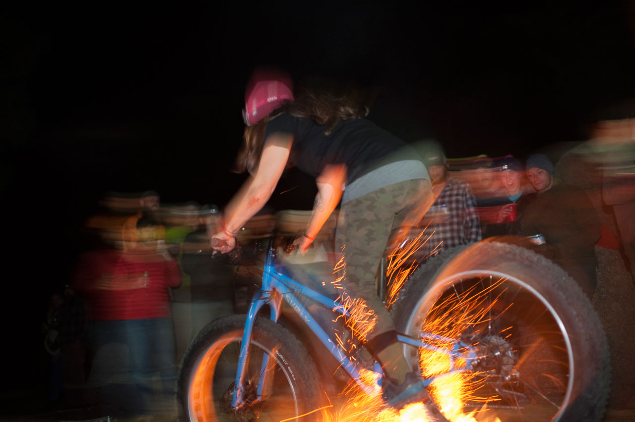 Left side, blurred view of a cyclist riding a blue Surly fat bike, over a campfire at night