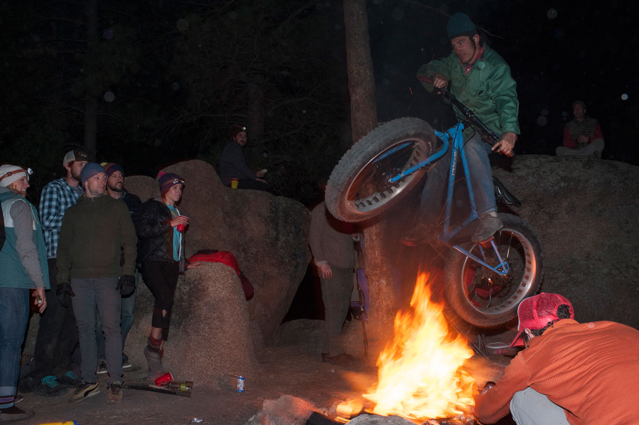 People gathered, watching a cyclist on a blue Surly fat bike, jumping over a campfire, at night in a forest