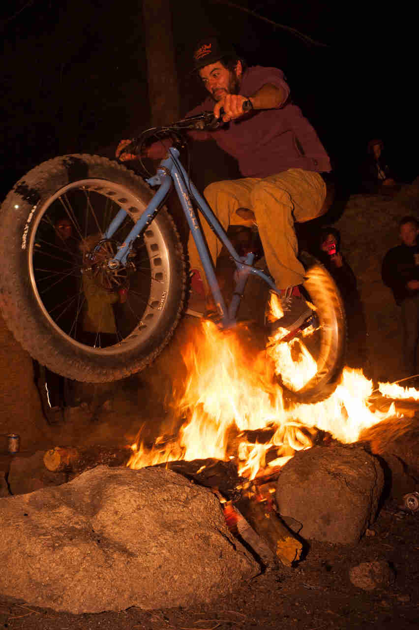 Front left side view of a cyclist riding a blue Surly fat bike, jumping over a campfire at night