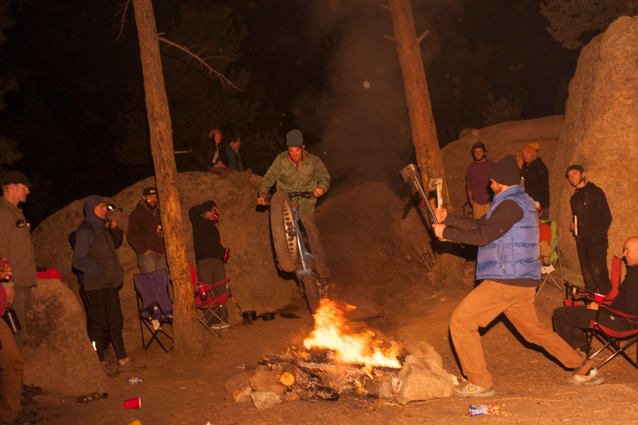 Front view of a cyclist on a Surly fat bike, jumping over a campfire at night, with spectators watching