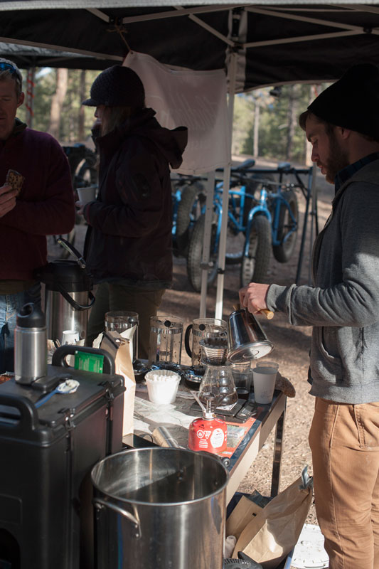 A person making a cup of coffee under a canopy, with people and fat bike in the background