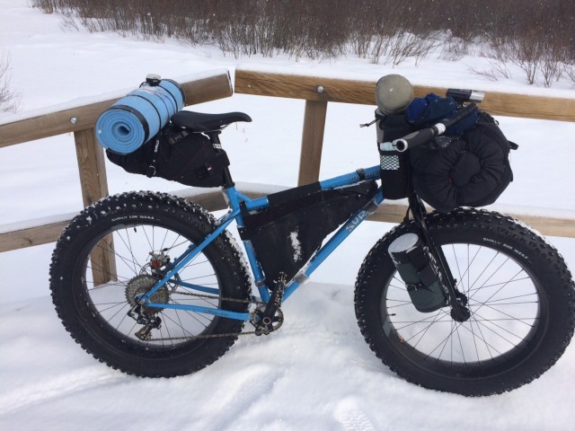 Right side view of a Surly Ice Cream Truck fat bike loaded with gear, standing in snow, with a wood post rail behind it