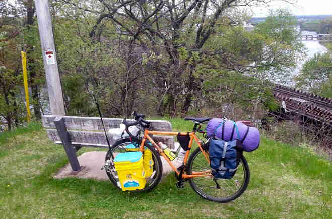 Left side view of an orange Surly bike, loaded with gear, parked on a grass hilltop, against the back or a park bench