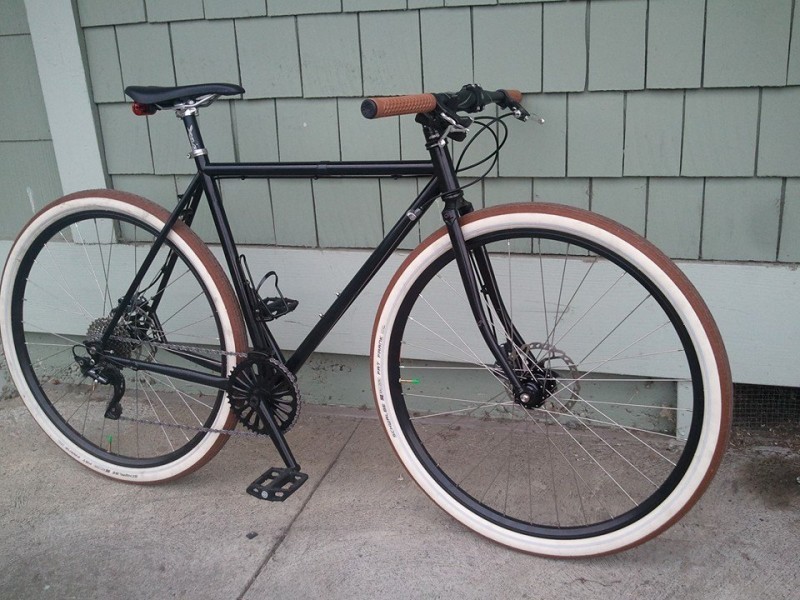 Right side view of a black Surly bike with white wall tires, parked on a sidewalk, leaning against a gray wall