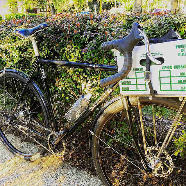 Right side view of a black Surly bike with cardboard box strapped to the front rack, parked against a wall of hedges