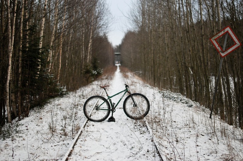 Right side view of a green Surly Krampus bike, standing across snowy railroad tracks, with bare trees on the sides
