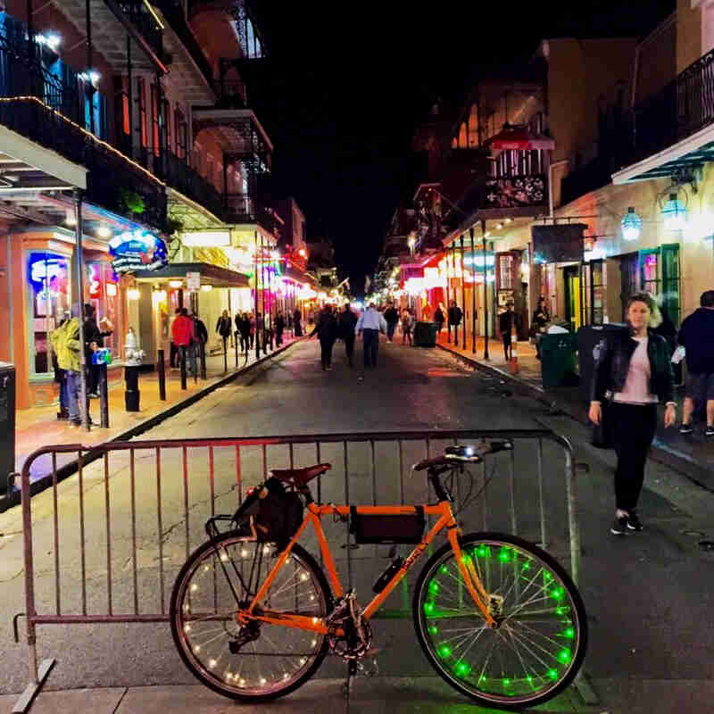 Right side view of an orange Surly bike with lighted spokes, parked in front of a gate, at Bourbon Street, at night