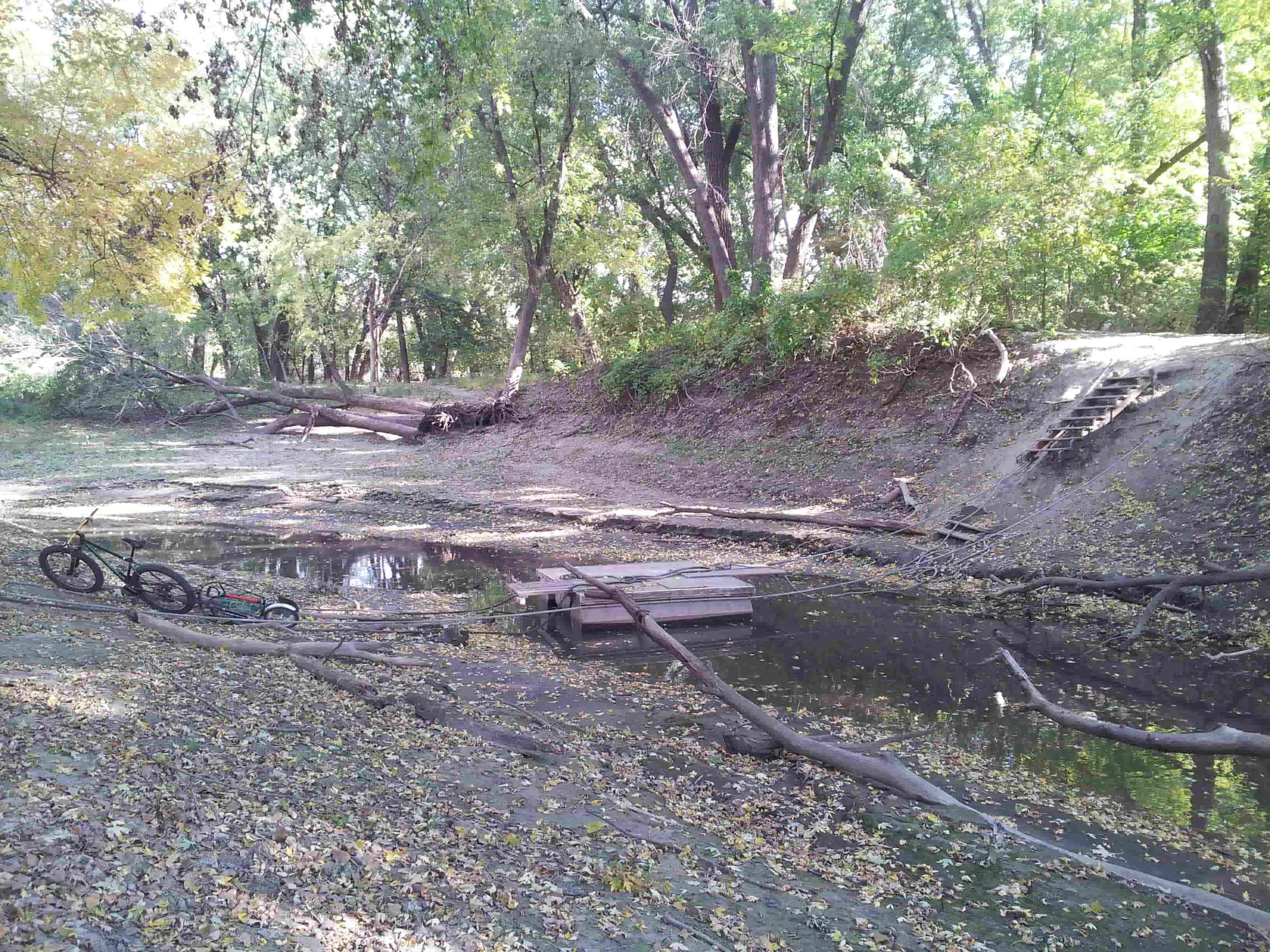 Left side view of a bike on a dirt bank, next to a river with a wood deck raft in the middle, in the green woods