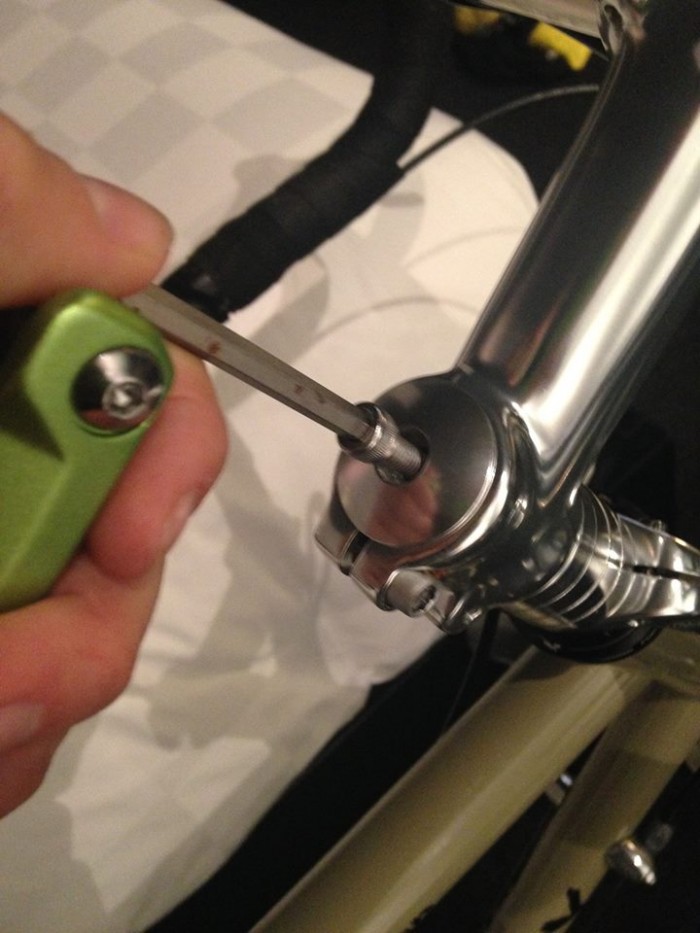 Downward, cropped view of a hand holding a hex wrench tool, removing the top bolt of a handlebar stem on a bike