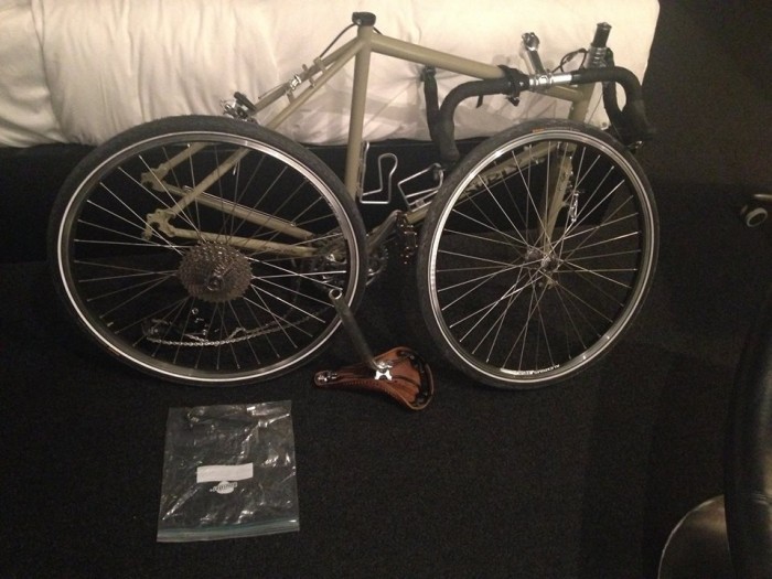 Right side view of a disassembled, tan Surly Long Haul Trucker bike, leaning against a bed in a room