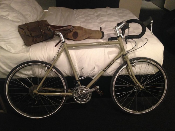 Right side view of a tan Surly Long Haul Trucker bike, parked against the side of a bed