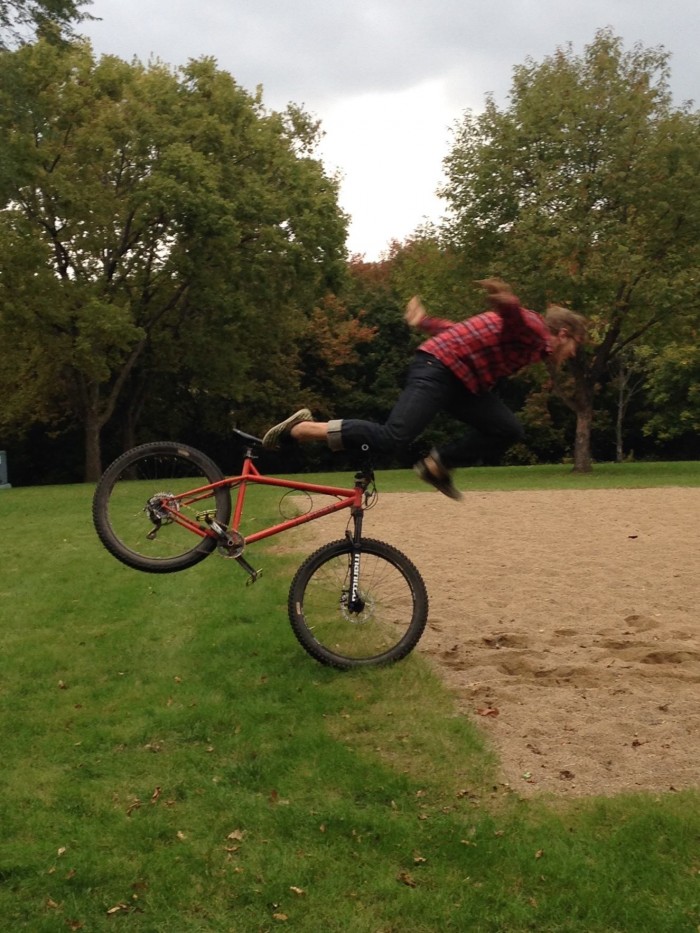 Right side view of a cyclist flying over the handlebars of a red bike, into a sand volleyball court below, at a park