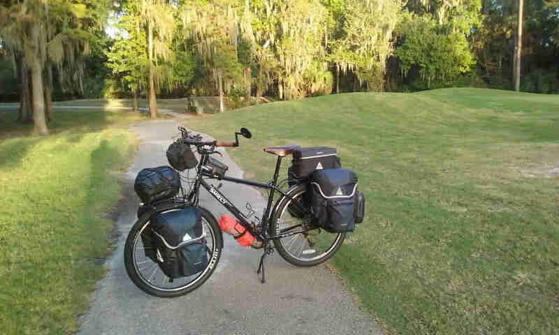 Left side view of a black Surly bike, loaded with gear packs, parked across a paved trail running through a green field