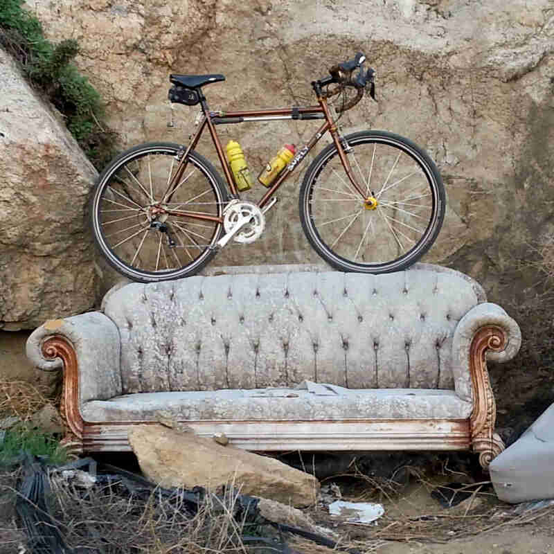 Right side view of a Surly Cross Check bike, parked on top of a couch backrest, sitting in dirt, in front of a rock wall