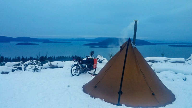 A teepee tent on a snow covered campsite, with a bike loaded with gear, behind, and a lake in the background