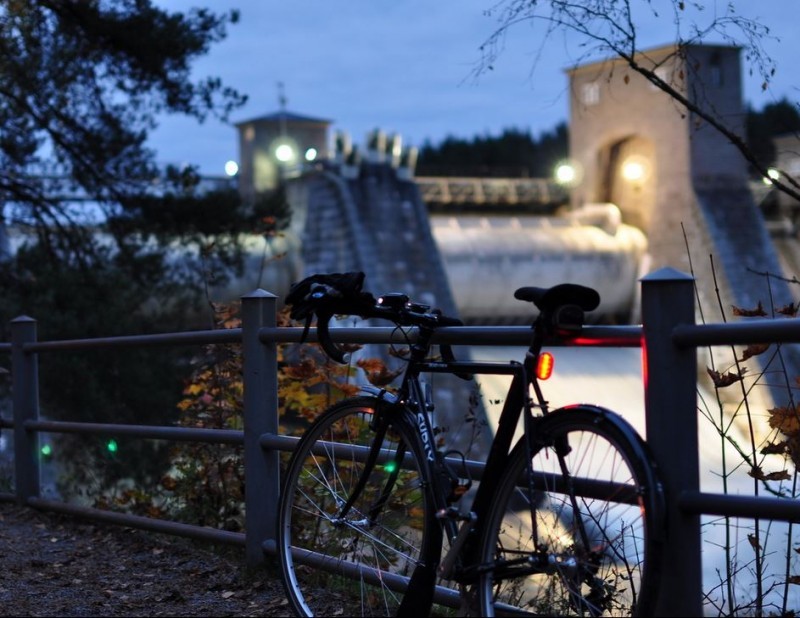 Rear, left side view of a Surly bike, parked against a steel handrail, with a large dam in the background, at nighttime
