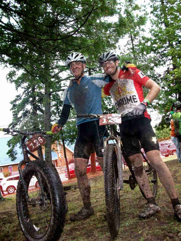 Two cyclists standing next to each other, covered in mud, with their dirty bikes